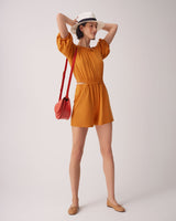 Woman in romper and hat holding her hat with a shoulder bag, standing.