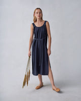 Woman in a sleeveless dress holding dried plants, standing against a plain background.