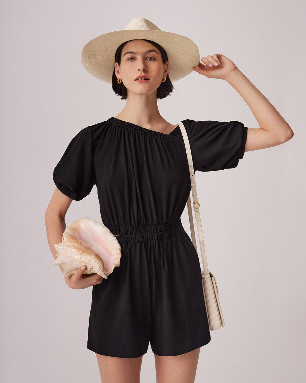 Woman in a hat and romper holding a shell, with a shoulder bag.