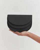 A hand holding a small, curved clutch bag against a plain background.