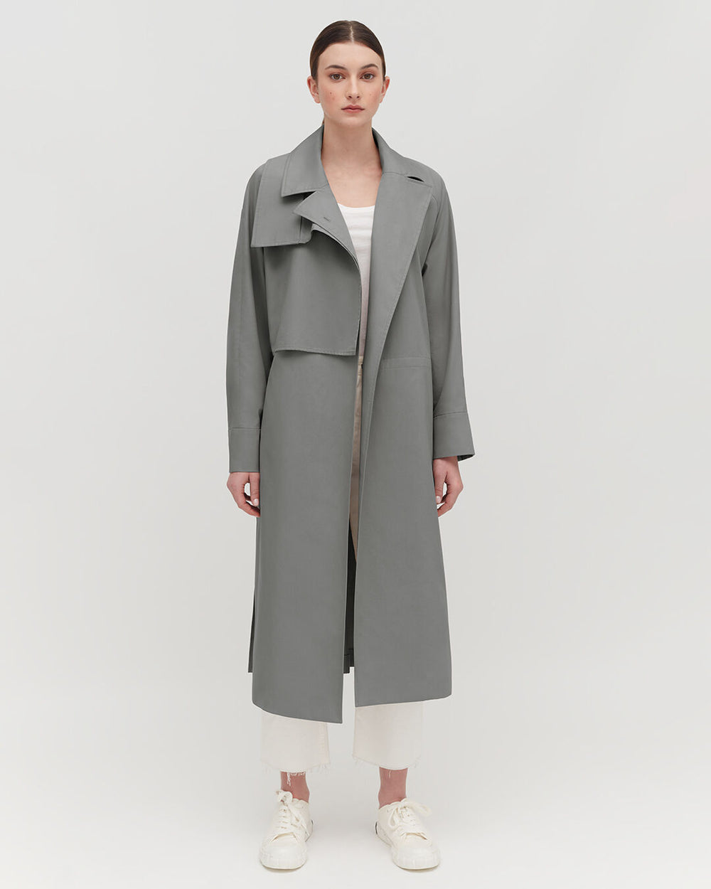 Woman in a long coat and sneakers standing against a plain background.
