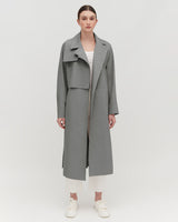 Woman in a long coat and sneakers standing against a plain background.