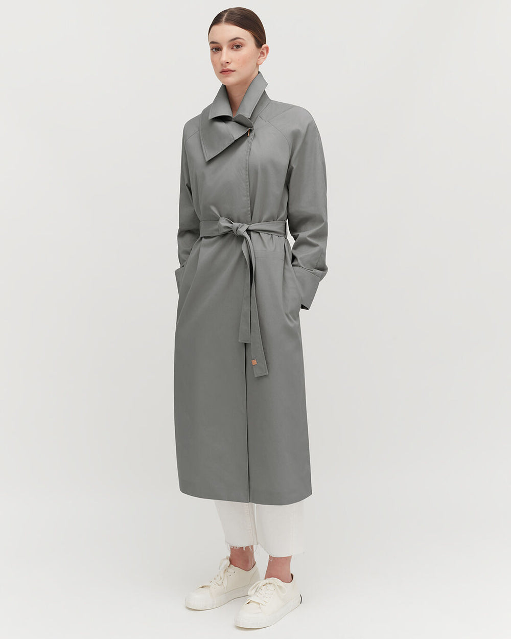 Woman standing in a coat with belt, wearing trousers and sneakers.