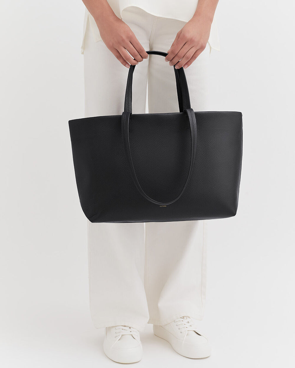 Person holding a large tote bag, standing in white pants and sneakers.