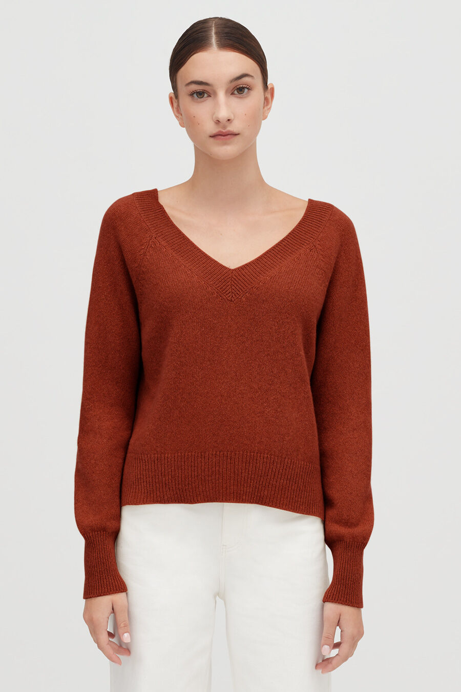 Woman in a v-neck sweater and white pants standing against a plain background.