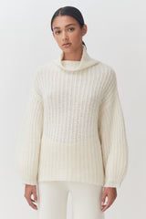 Woman in turtleneck sweater and pants standing against plain background.