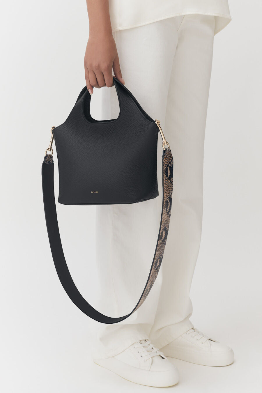 Person holding a handbag with a strap, standing in pants and shoes.