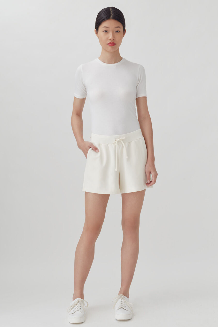Woman standing in a plain background wearing a short-sleeve top and shorts with sneakers.