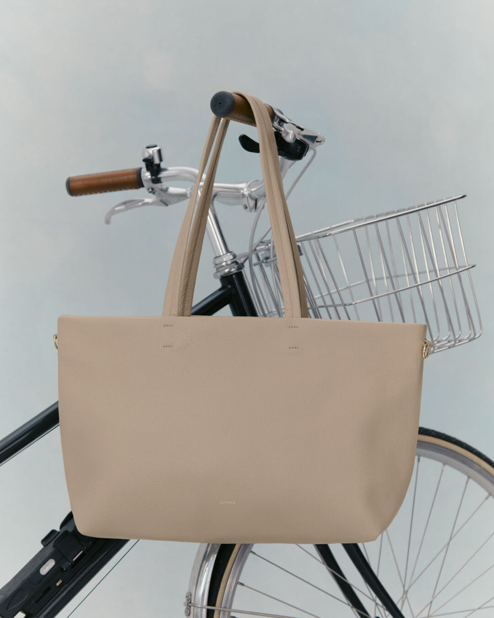 Handbag hanging on bicycle handlebar with a basket in the background.