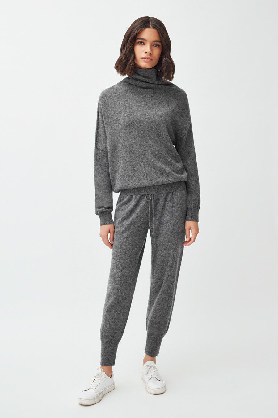 Woman standing in turtleneck and pants with hands in pockets.