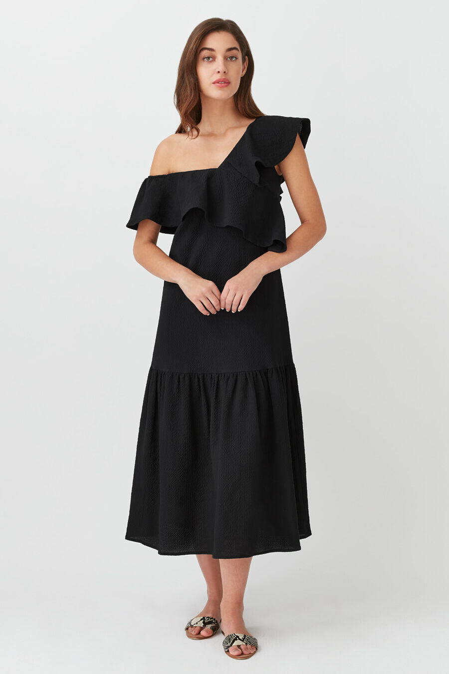 Woman in dress with one shoulder strap and ruffled layers, standing straight.