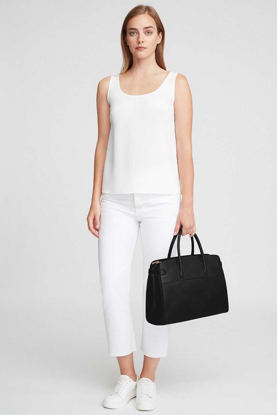 Woman standing holding a handbag, wearing a sleeveless top and pants with sneakers.
