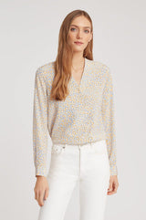 Woman standing wearing a patterned blouse and white pants.